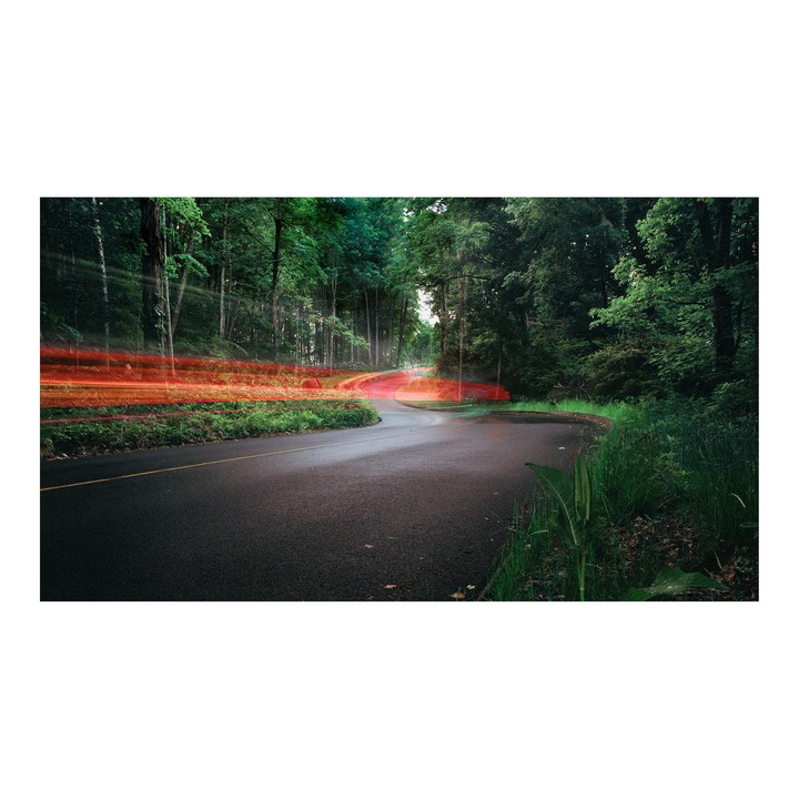 A winding road through a forest shows red light trails from a vehicle at night.