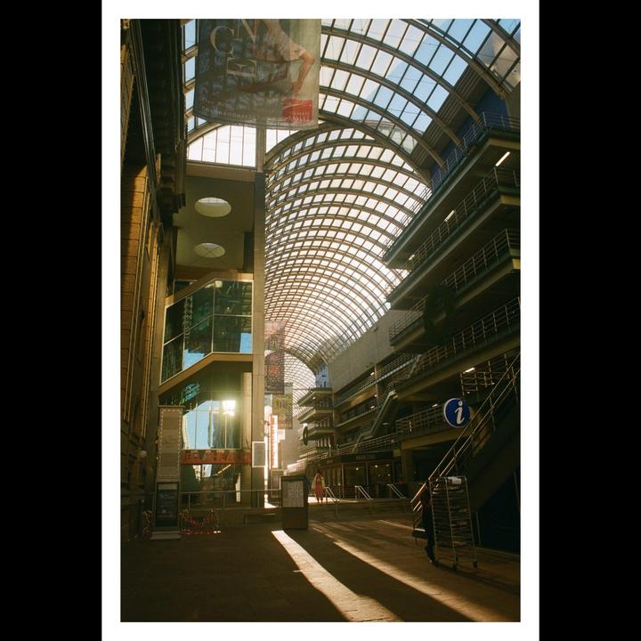 Sunlight streams through the glass arched roof of a large indoor space with multiple levels of balconies and a person walking in the distance.
