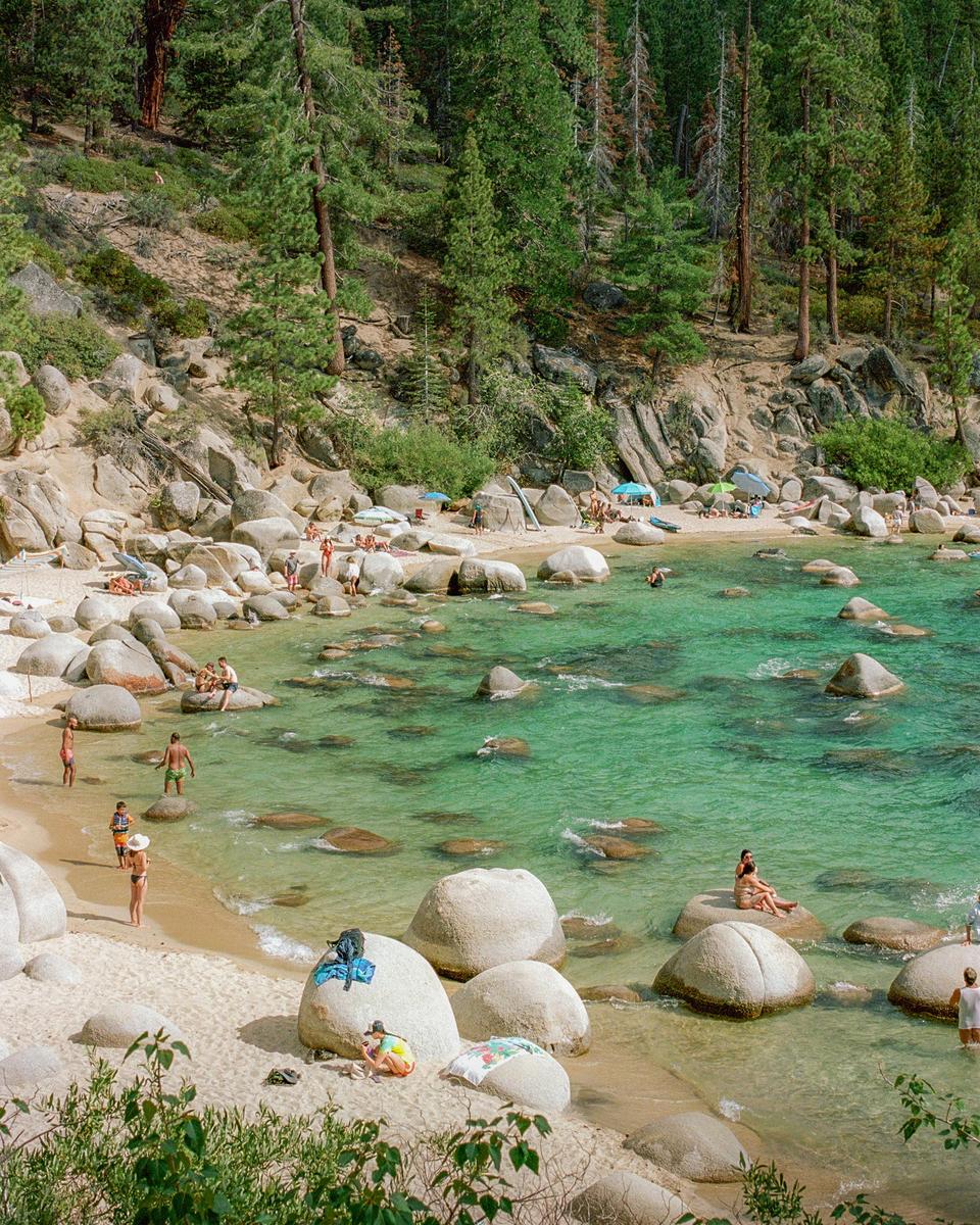 A sandy beach with large boulders and clear turquoise water is filled with people swimming and sunbathing, with a dense forest in the background.