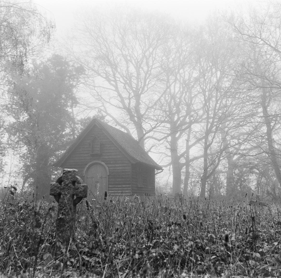 A small wooden chapel stands in a field of tall grass with a stone cross in the foreground, all shrouded in fog.