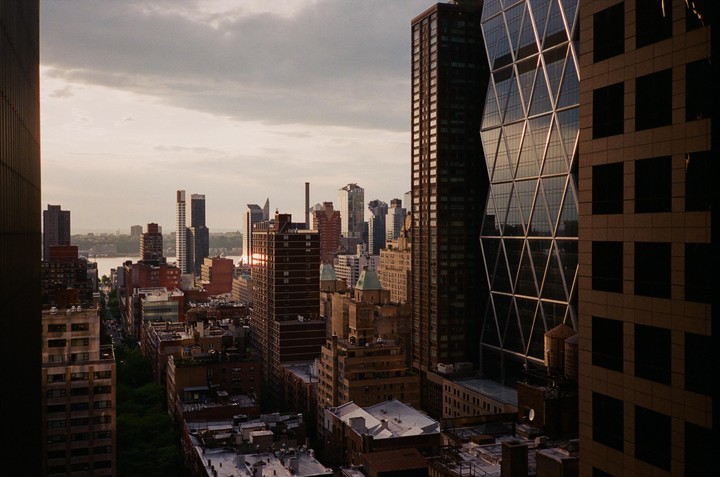 A cityscape with tall buildings and a river in the distance under a cloudy sky.