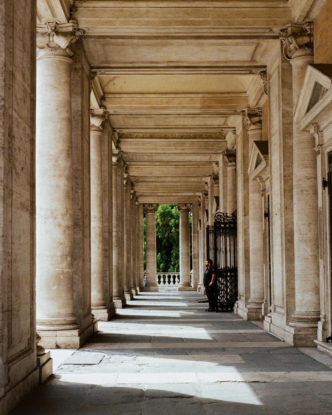 A man stands in a long, columned hallway with light and shadow patterns on the floor.