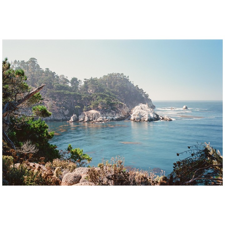 A hazy coastal landscape shows a tree-covered peninsula jutting into a calm blue ocean with white rock formations along the shore.
