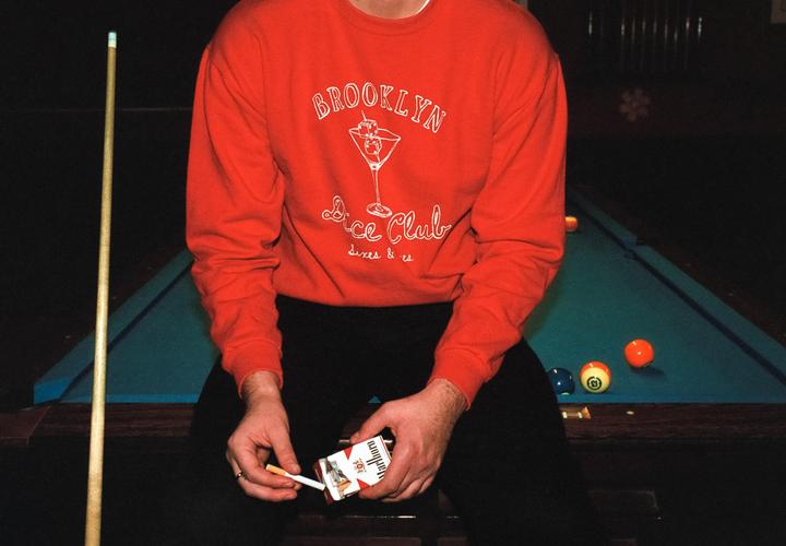 A man in a red sweatshirt sits next to a pool table with a cigarette in his hand.
