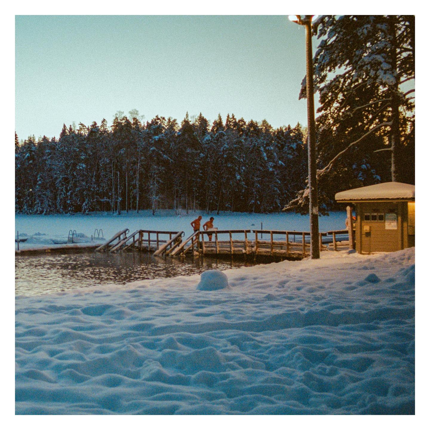 Two people stand on a wooden pier over a lake surrounded by snow-covered ground and trees.