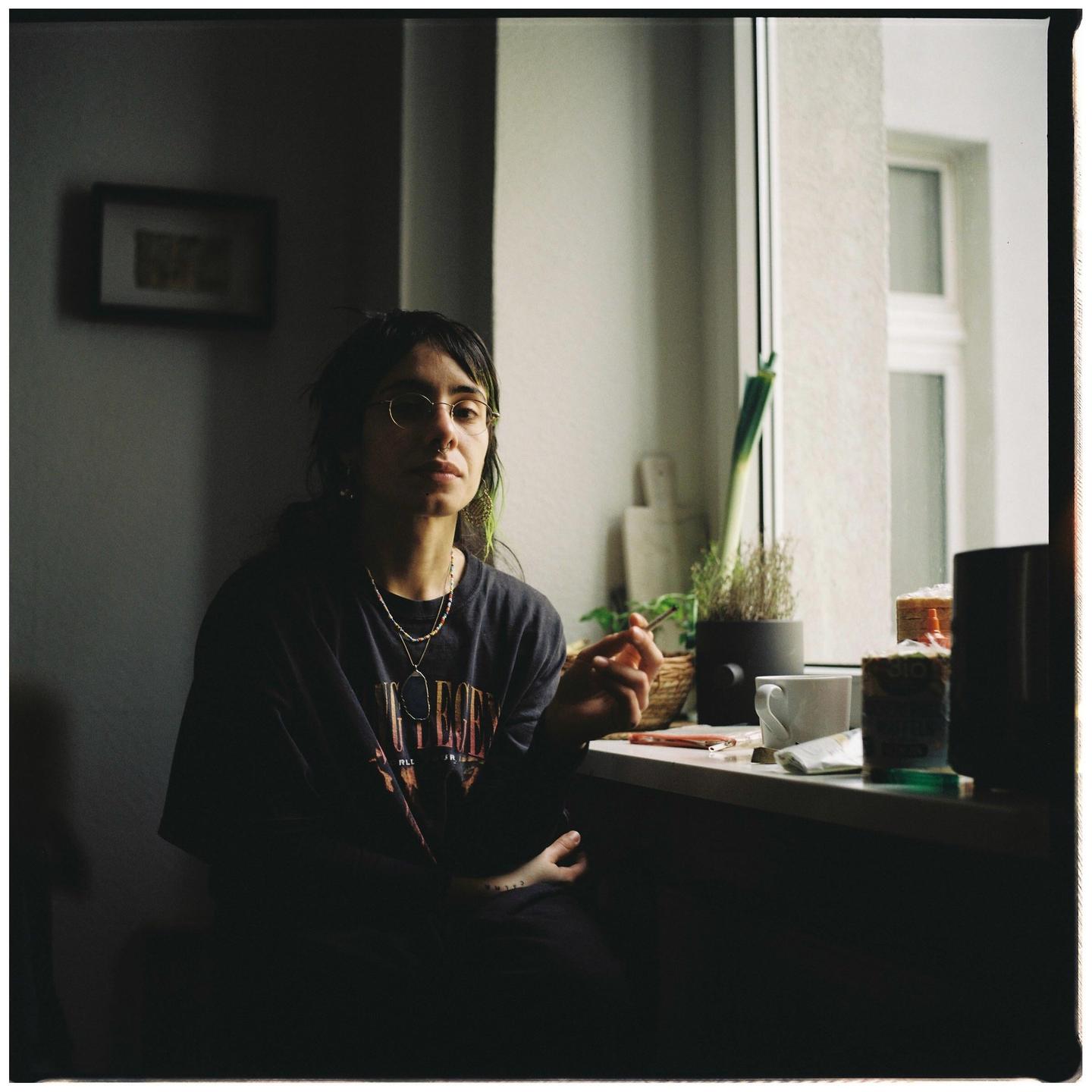 A woman with glasses sits by a window, holding a small stick.