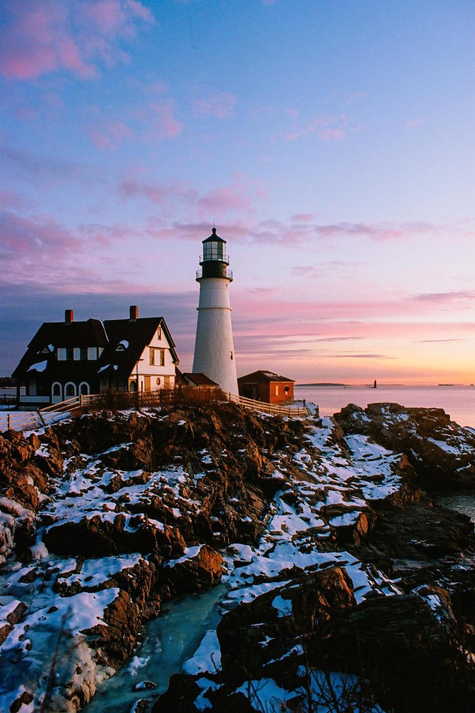 A lighthouse and a house stand on a rocky, snow-covered coast under a colorful sky at sunrise.
