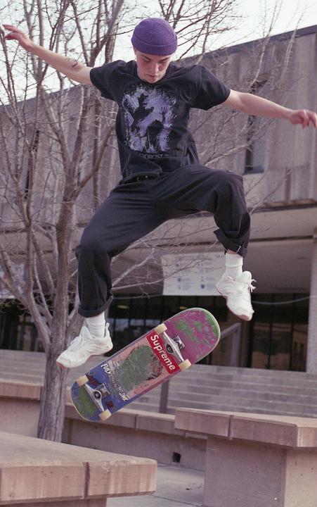A young man in a purple beanie and black t-shirt jumps with a skateboard in mid-air.