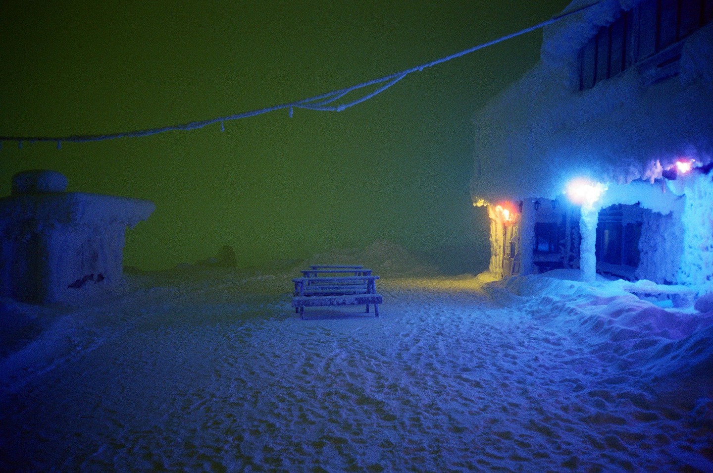 A snow-covered building with blue and orange lights illuminates a picnic table and the surrounding snow under a green sky.
