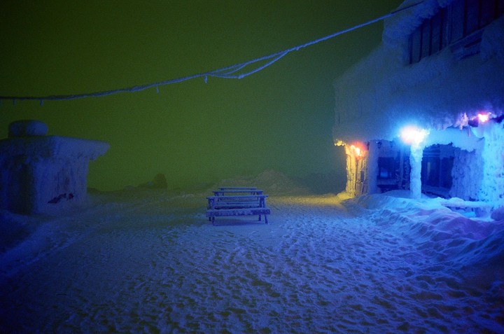 A snow-covered building with blue and orange lights illuminates a picnic table and the surrounding snow under a green sky.
