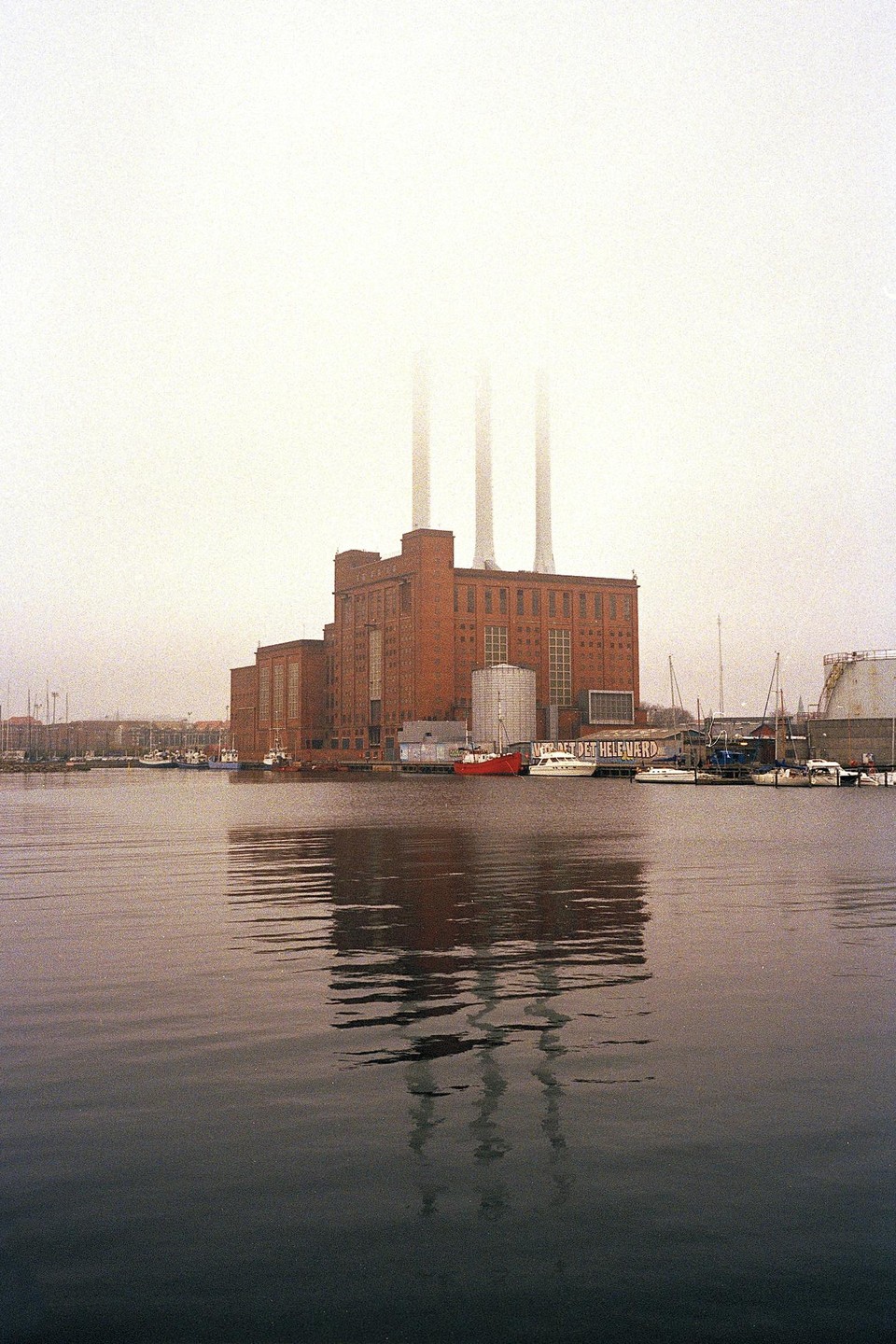 A large brick power plant with three tall smokestacks is reflected in the water of a harbor filled with boats on a foggy day.
