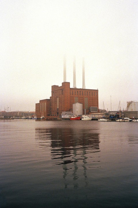 A large brick power plant with three tall smokestacks is reflected in the water of a harbor filled with boats on a foggy day.