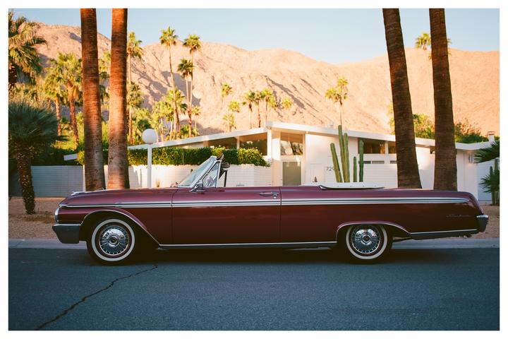 A red convertible car is parked on a street in front of a white building with palm trees and mountains in the background.