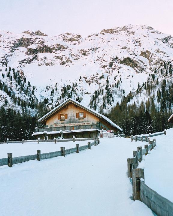 A wooden cabin with a fence in front of it is surrounded by snow-covered mountains and pine trees.