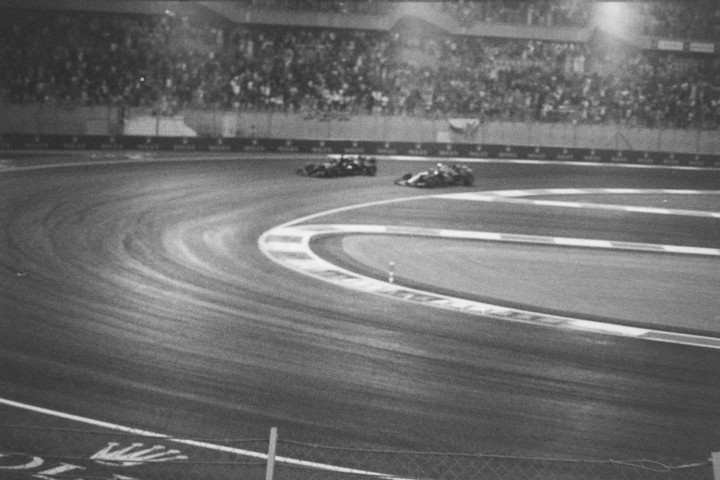 Two race cars speed around a track at night with a crowd visible in the background.
