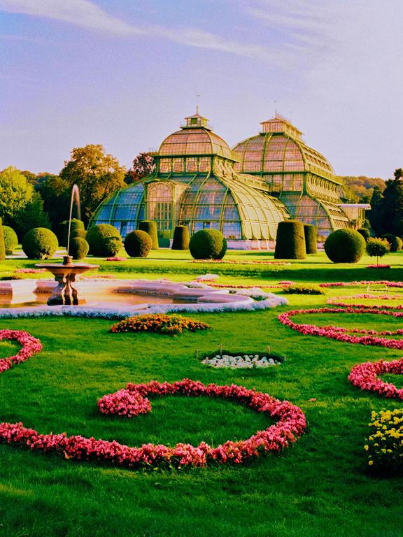 A large glass greenhouse stands behind a manicured garden with a fountain and flower beds.