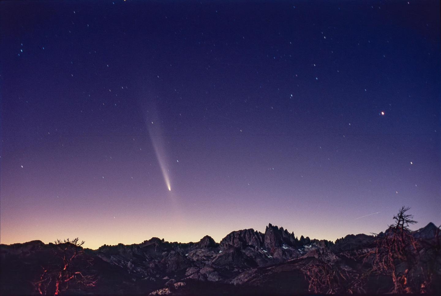 A comet with a bright tail is visible in the night sky above a mountain range.