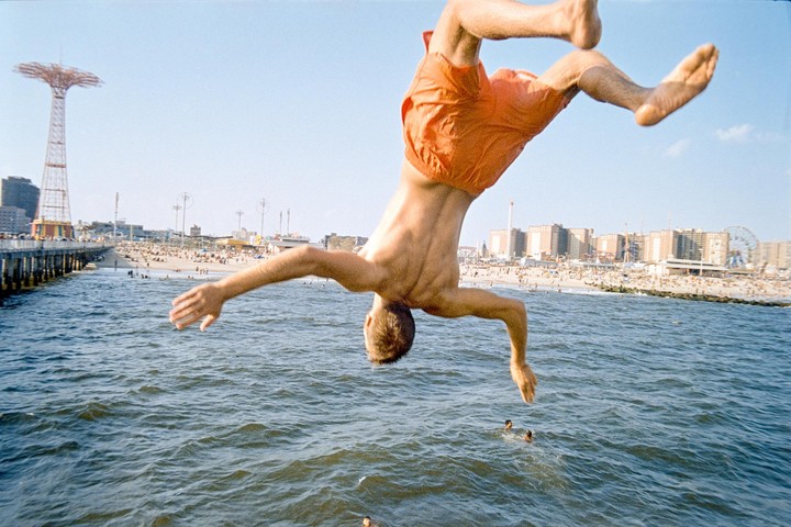 A man in orange swim trunks dives headfirst into the ocean from a pier, with a crowded beach and amusement park in the background.