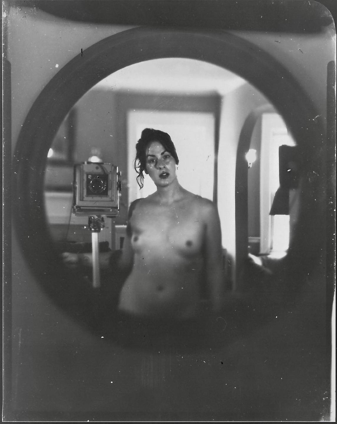 A topless woman is reflected in a round mirror, with a large format camera visible to her left.
