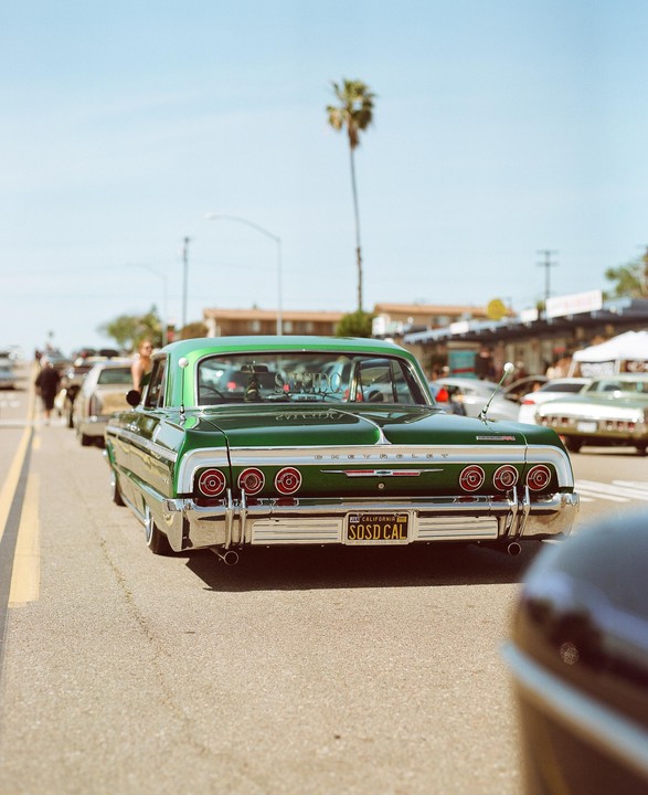 A green Chevrolet Impala is parked on a street with a palm tree in the background.