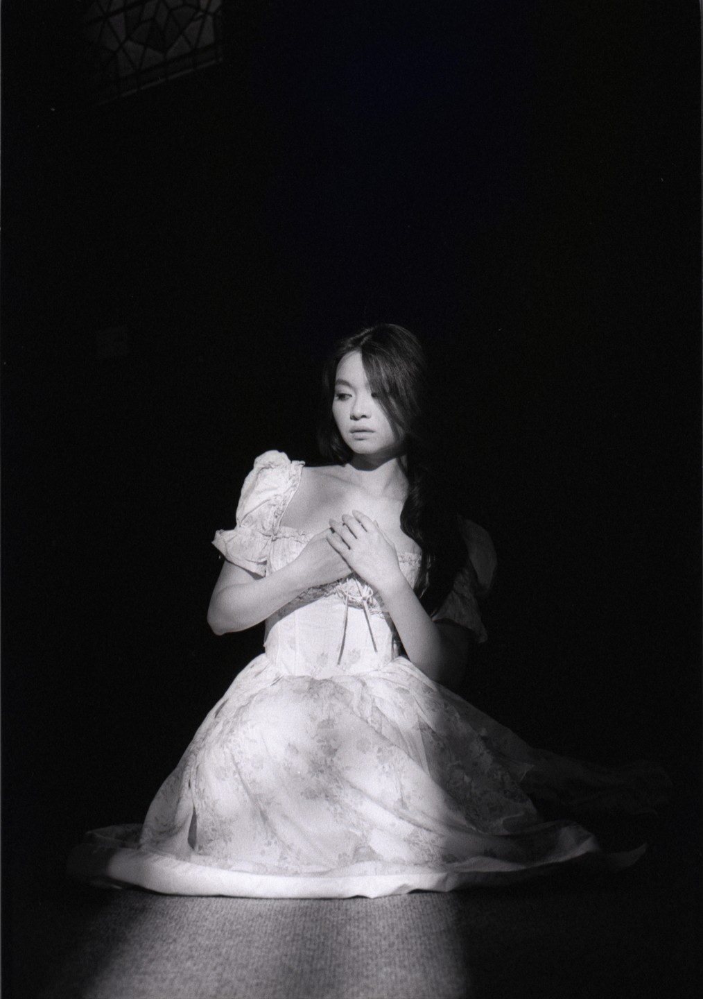 A woman in a white dress kneels on a dark floor, illuminated by a spotlight from above.