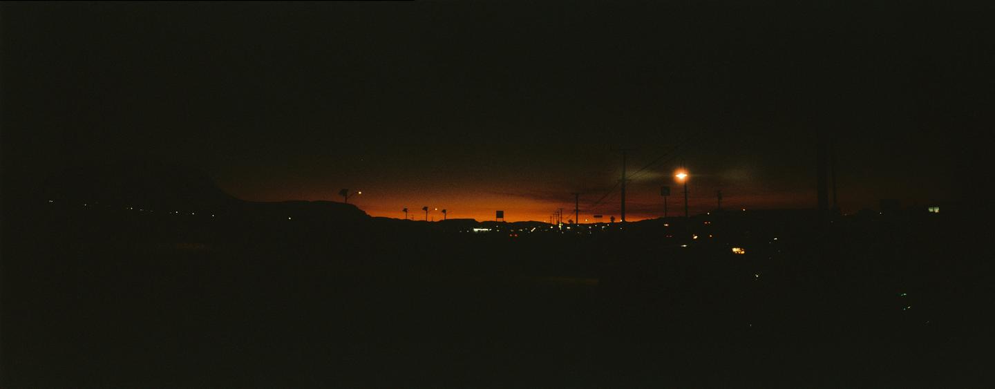 A panoramic view of a city at dusk shows a bright orange sky above a dark landscape with scattered lights.