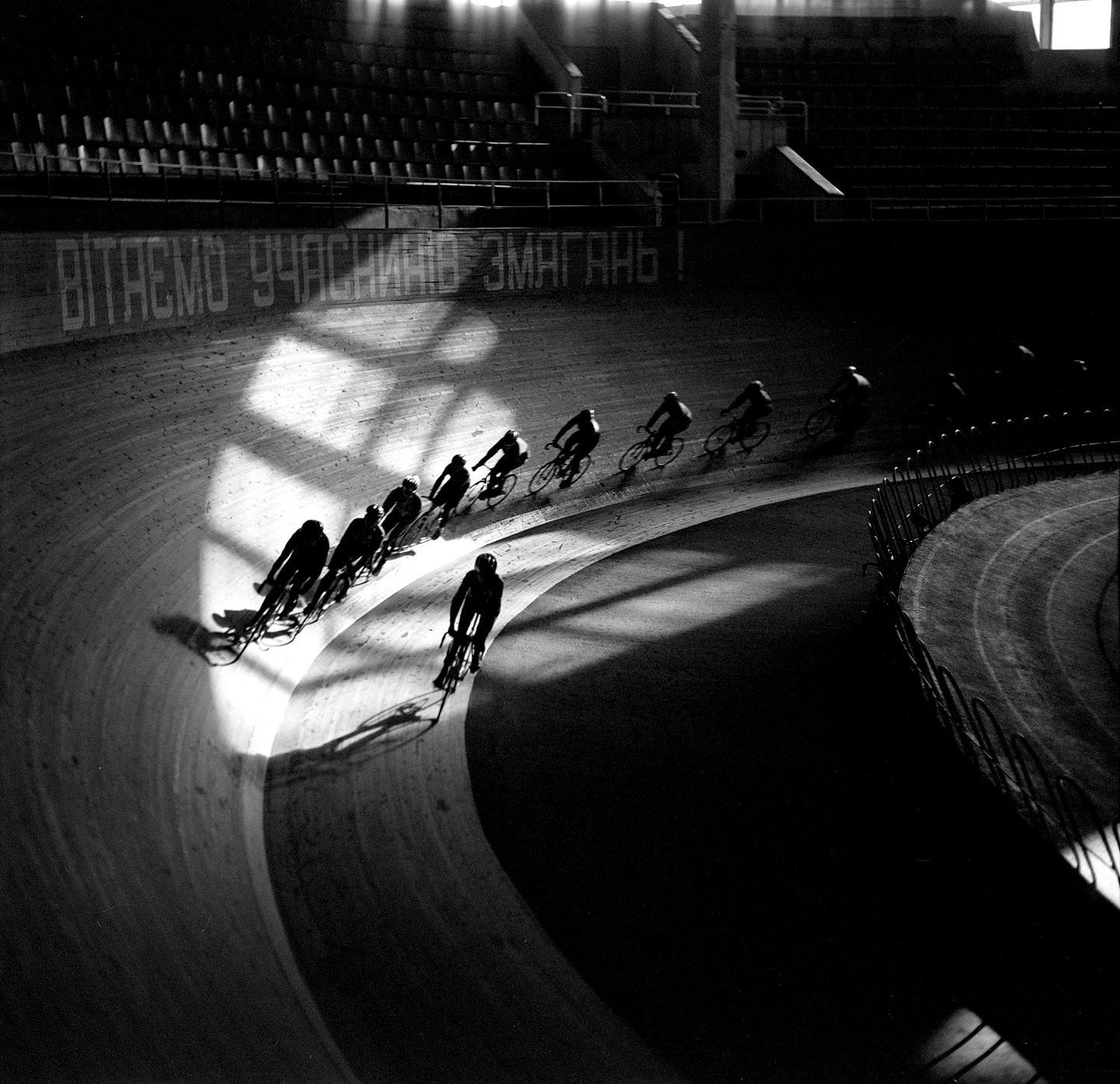 A group of cyclists ride along a velodrome track with strong shadows and light.