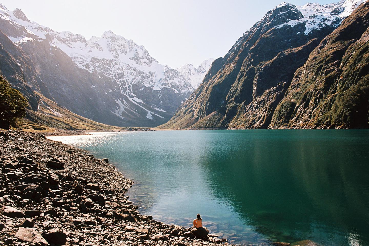 A person sits on a rock at the edge of a lake surrounded by mountains.