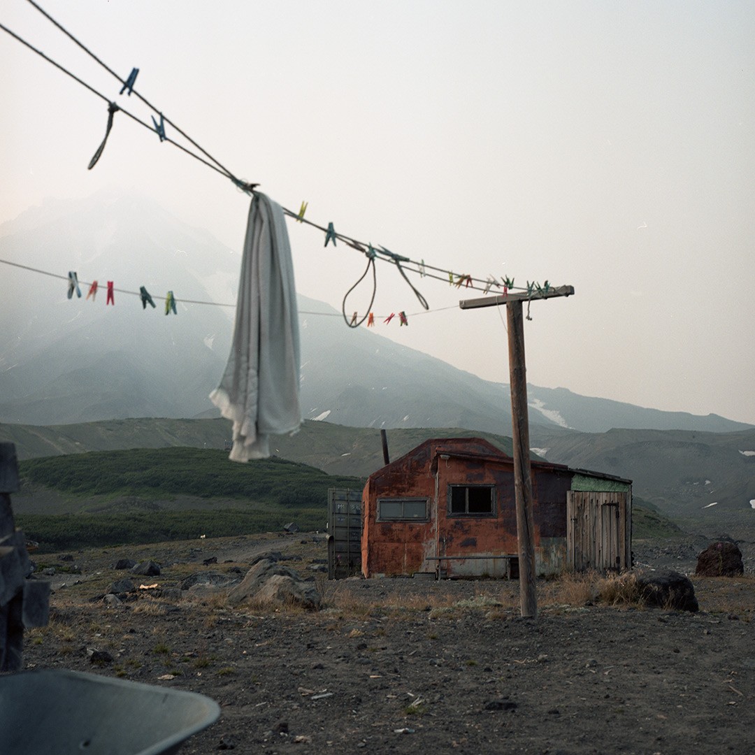 A clothesline with a white sheet and clothespins hangs in front of a small, rusty hut with a mountain in the background.