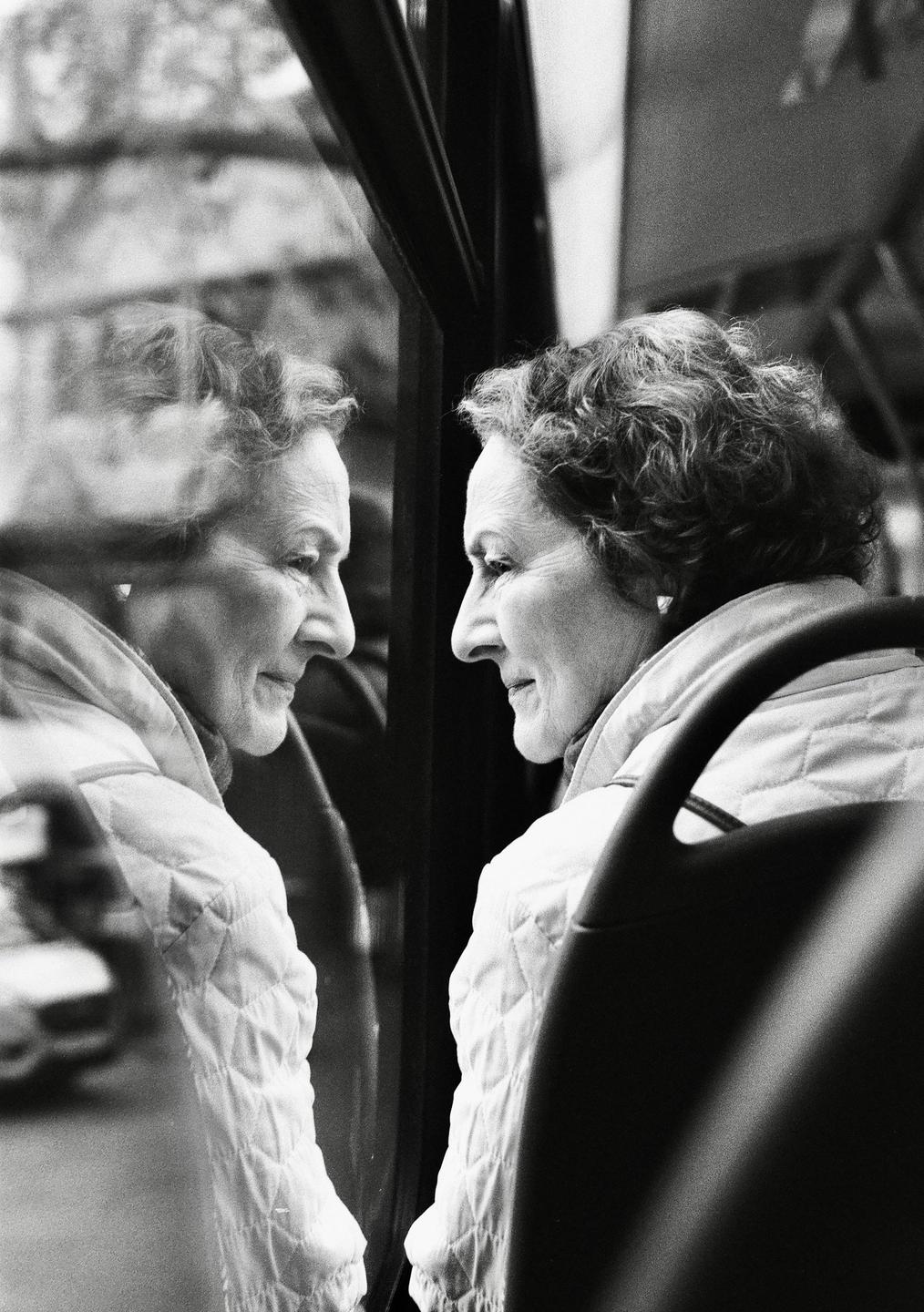 A woman with curly hair looks out a window, her reflection visible beside her.