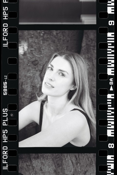 A woman with long hair looks towards the camera, framed by the sprocket holes and edge markings of a film strip.