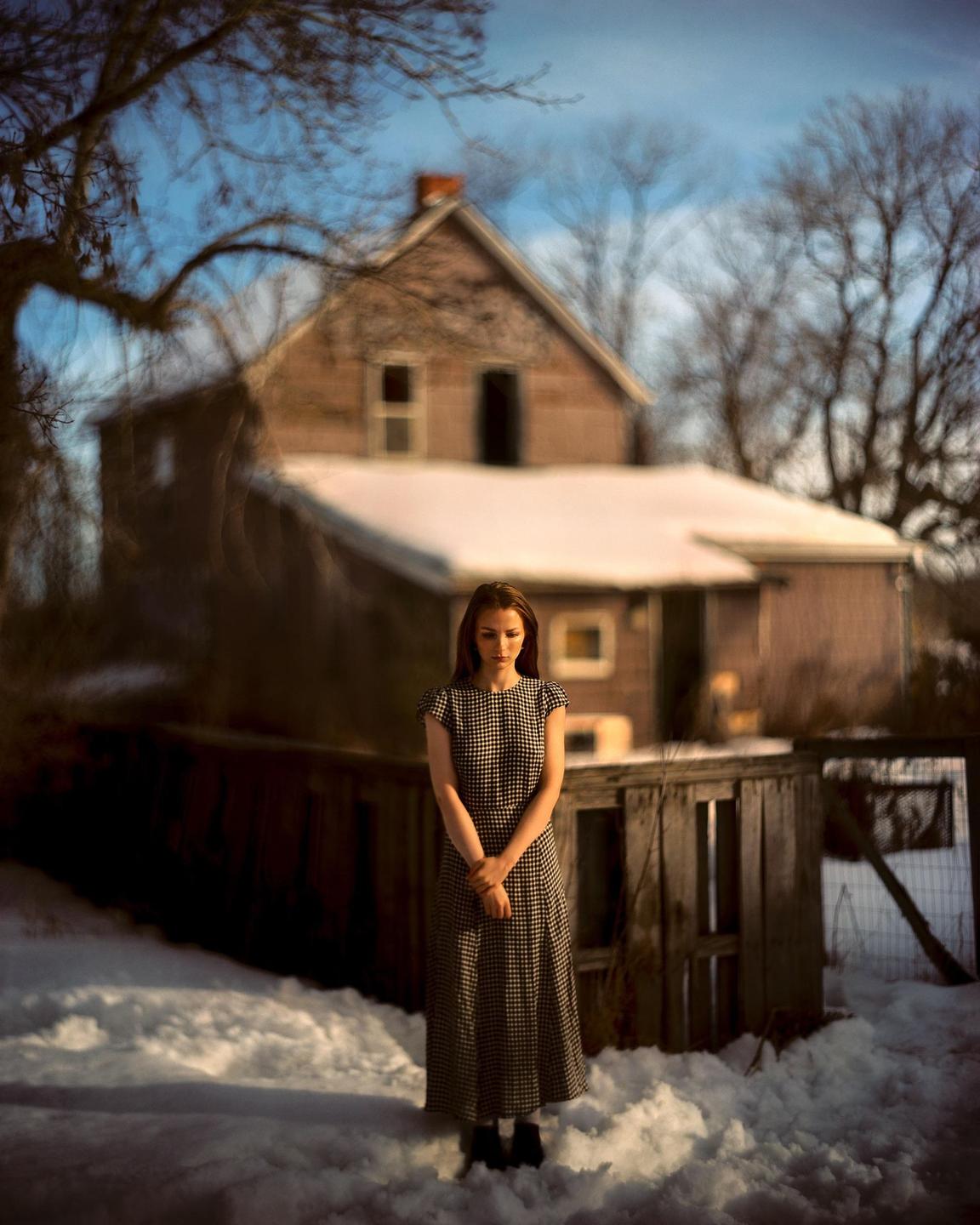 A woman in a long dress stands in the snow in front of a house.
