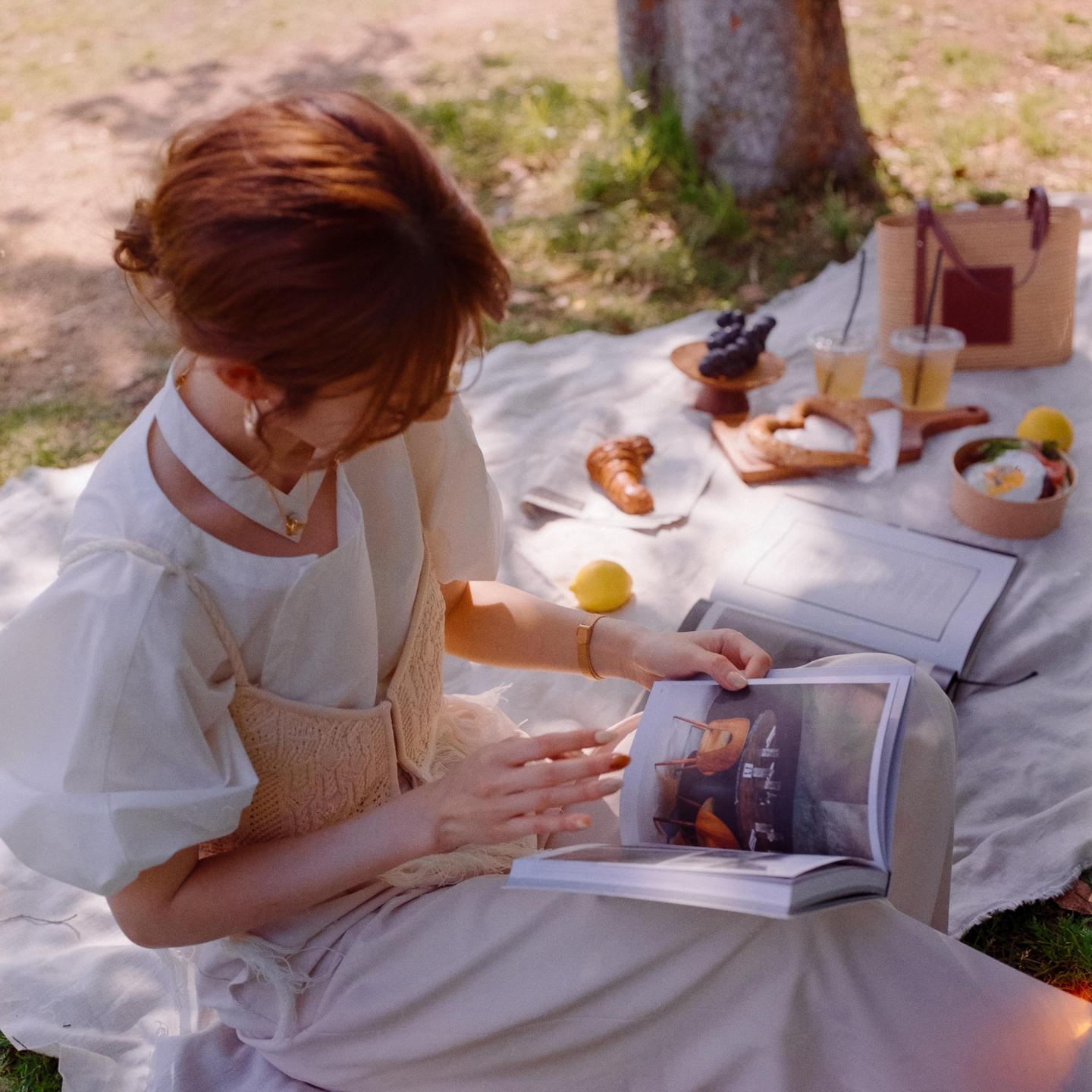 A woman sits on a blanket on the grass, reading a book with a picnic spread next to her.