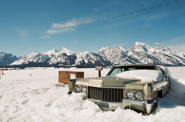 A vintage car is partially buried in snow with a mountain range in the background under a clear blue sky.
