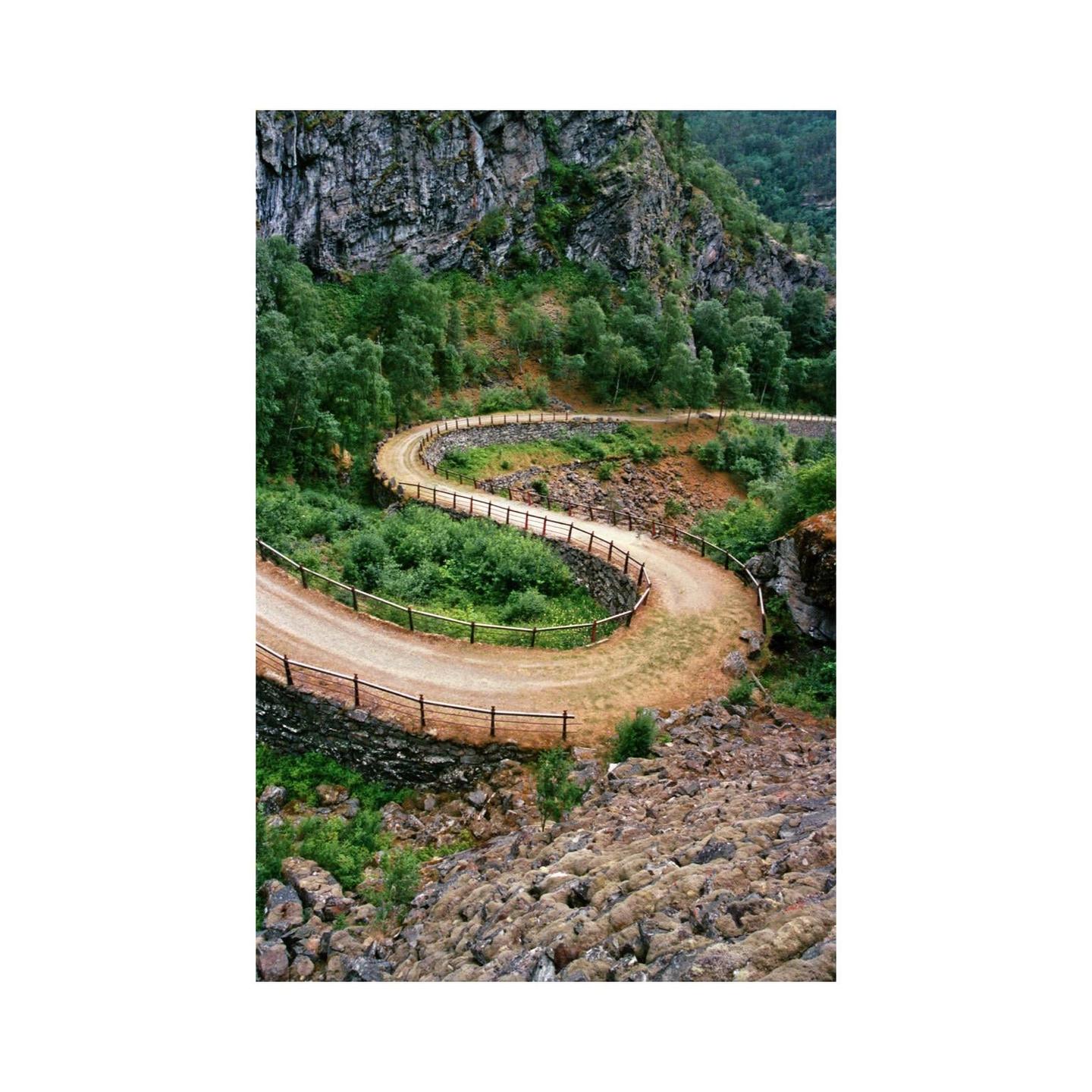 A winding dirt road with a wooden fence curves through a lush green valley with a rocky mountain face in the background.