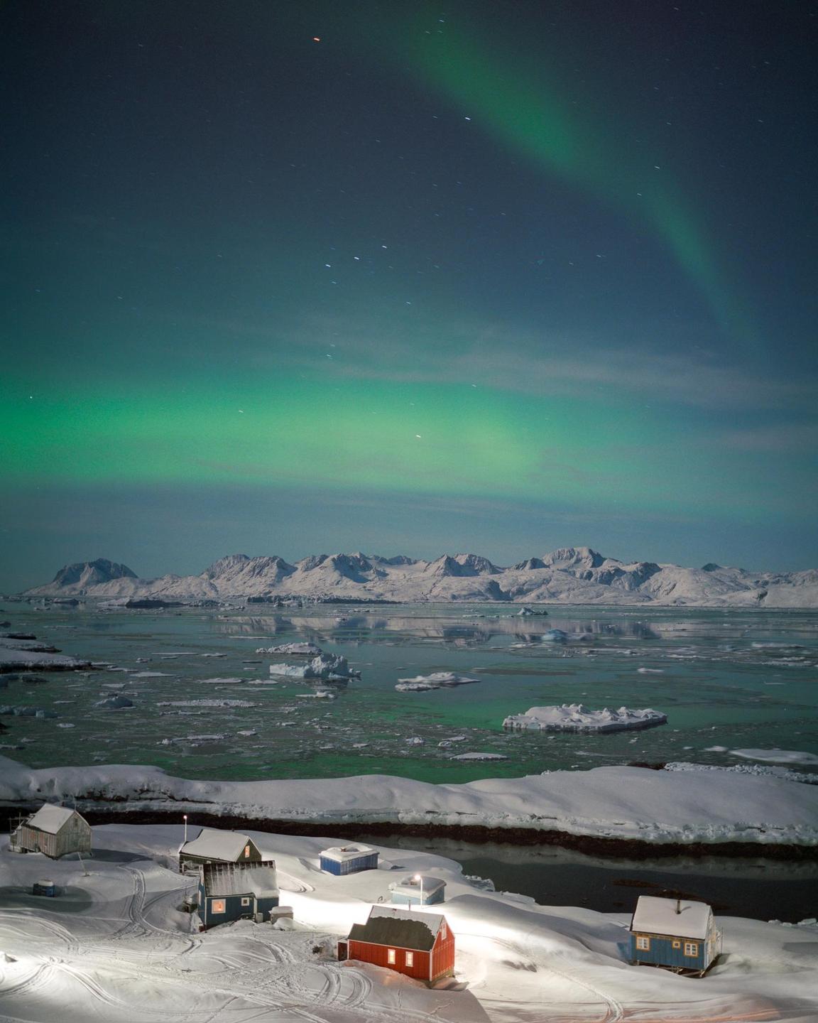 A small village with colorful houses sits on a snow-covered shore under a vibrant aurora borealis and a starry night sky, with mountains and ice-filled water in the distance.