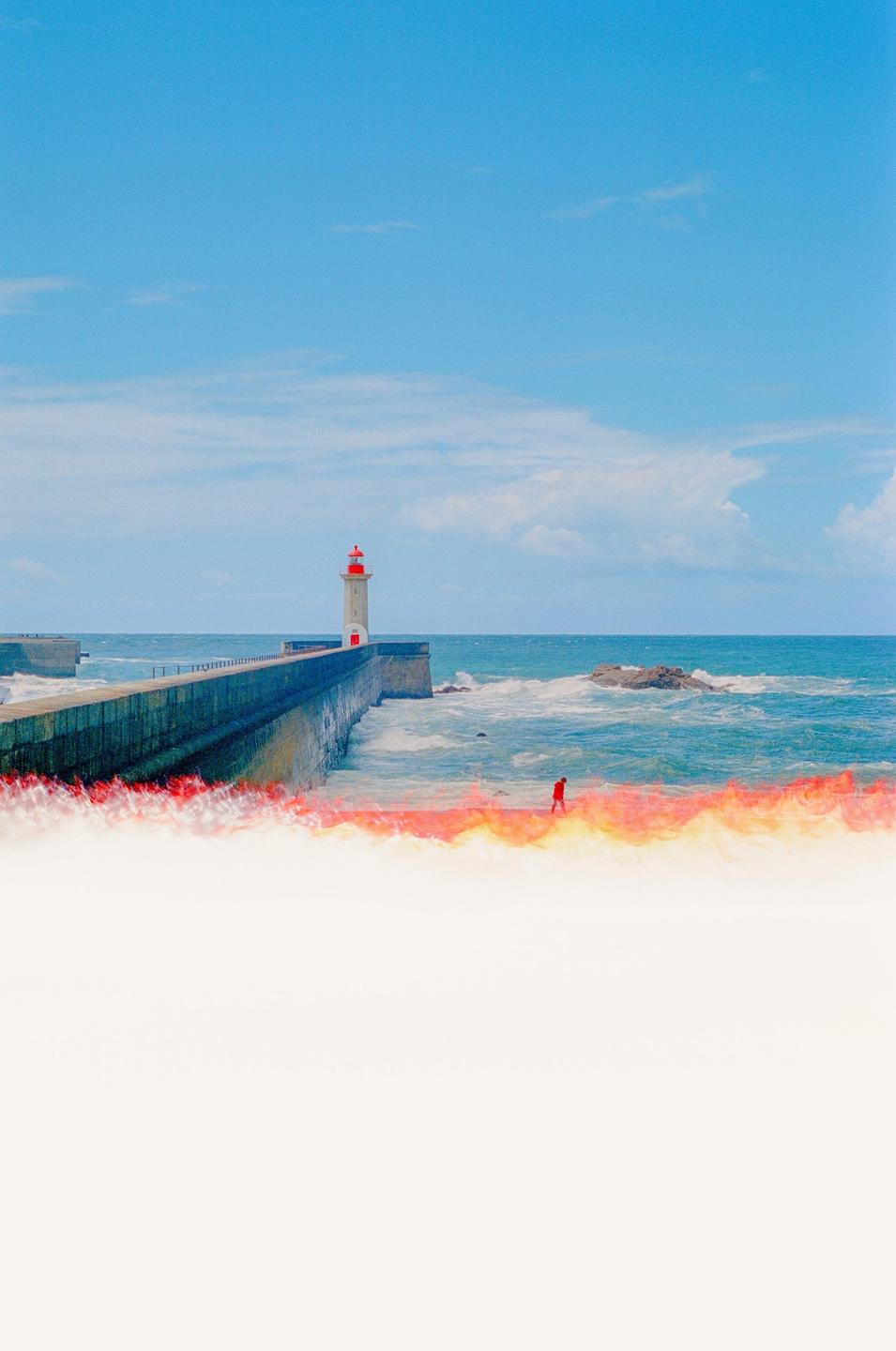 A lighthouse stands at the end of a long pier with a person walking along the water's edge.