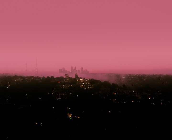 A city skyline is visible through a thick layer of fog under a vibrant pink sky.