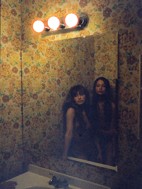 A woman and her reflection are visible in a mirror above a sink in a room with floral wallpaper.