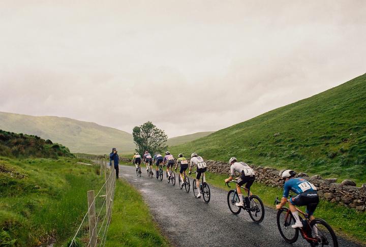 A group of cyclists ride along a wet road through a green, hilly landscape with a person watching from the side.