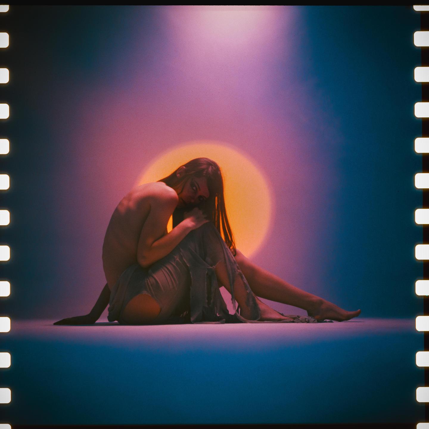 A woman sits on the floor with a bright orange circle behind her.