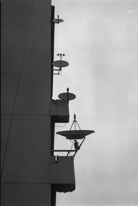 Three satellite dishes are mounted on the side of a building against a bright sky.