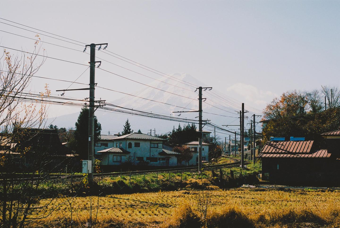 A snow-capped mountain rises above a rural landscape with houses, power lines, and a railway track.