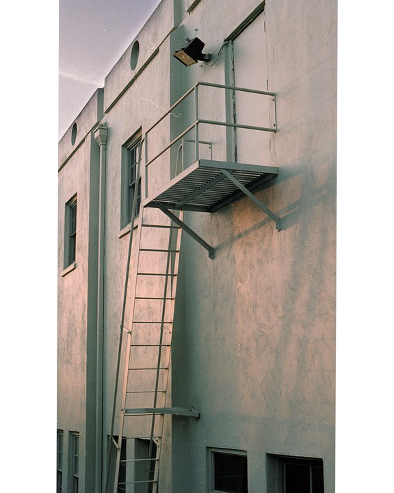 A metal fire escape ladder and platform are attached to the side of a light-colored building with windows.