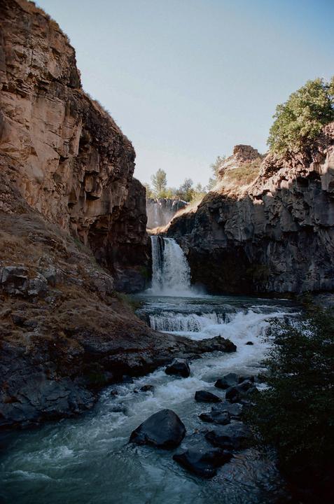 A waterfall cascades into a river surrounded by rocky cliffs and sparse vegetation under a clear sky.