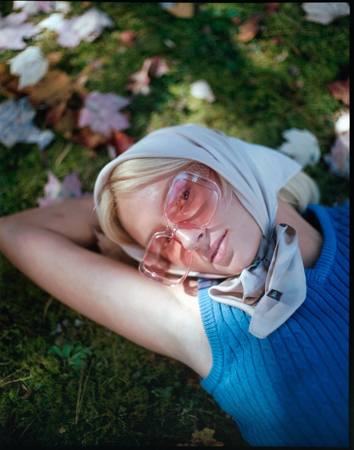 A woman wearing a headscarf and pink sunglasses lies on grass with autumn leaves.