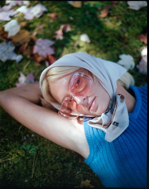A woman wearing a headscarf and pink sunglasses lies on grass with autumn leaves.