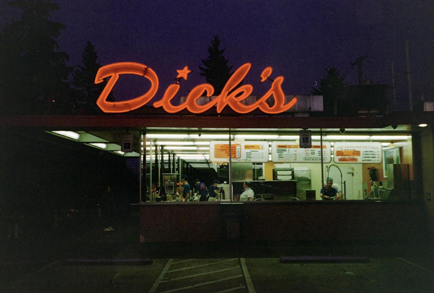 A neon sign for Dick's restaurant glows orange at night above the brightly lit interior of the establishment where several people are working.