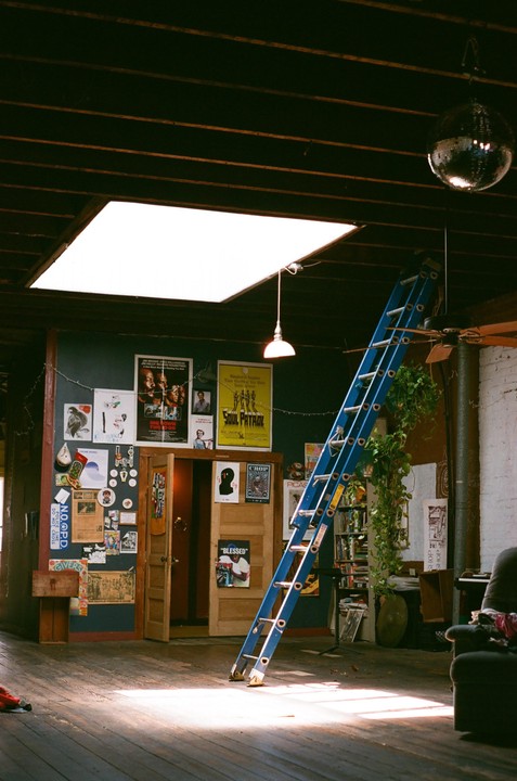 A blue ladder leans against a wall covered in posters and stickers in a room with a skylight and a disco ball.