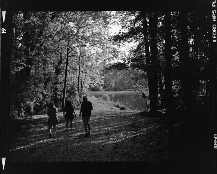Three people walk along a path next to a lake surrounded by trees.